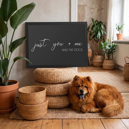 A quote print (Charcoal / Black Frame) reading You Me and the Dogs, in a boho living room with rattan, plants and a jute rug, with Simba nearby. [ks Background=Charcoal; Style=Black Frame; kind=lifestyle]