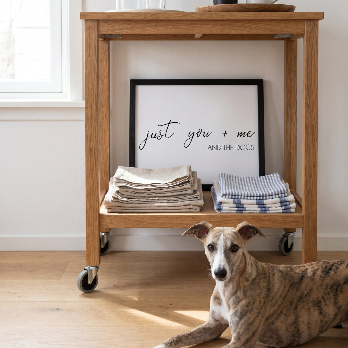 A quote print (White / Black Frame) reading You Me and the Dogs, in a sunny Scandinavian bedroom with white walls and oak furniture, with Arrow nearby. [ks Background=White; Style=Black Frame; kind=lifestyle]