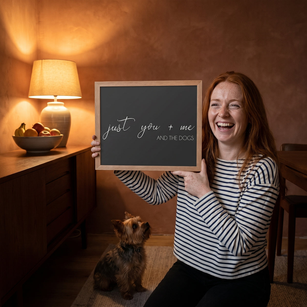 A red-haired freckled woman in her 30s in a striped Breton top with a quote print (Charcoal / Oak Frame) reading You Me and the Dogs, with Gizmo beside them. [ks Background=Charcoal; Style=Oak Frame; kind=lifestyle]
