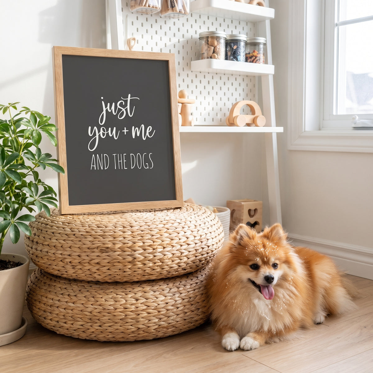 A quote print (Charcoal / Oak Frame) reading You Me and the Dogs, in a light-filled nursery-turned-craft room with pegboard shelves, with Nugget nearby. [ks Background=Charcoal; Style=Oak Frame; kind=lifestyle]