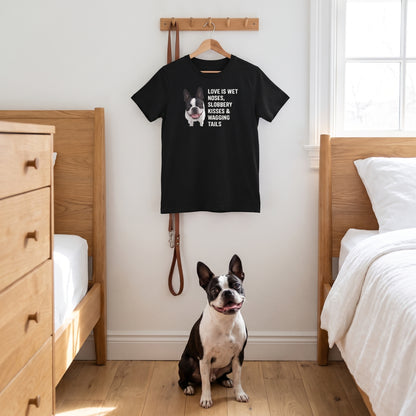 A custom pet t-shirt (Black) printed with "Love Is Wet Noses, Slobbery Kisses & Wagging Tails" and an illustration of Ziggy the Boston Terrier, in a sunny Scandinavian bedroom with white walls and oak furniture, with Ziggy nearby. [ks Colour=Black; kind=lifestyle]