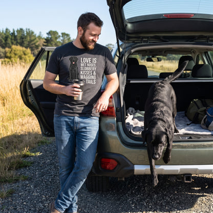A bearded man in his early 30s wearing a custom pet t-shirt (Dark Grey Heather) printed with "Love Is Wet Noses, Slobbery Kisses & Wagging Tails" and an illustration of Bella the black Labrador, with Bella beside them. [ks Colour=Dark Grey Heather; kind=lifestyle]