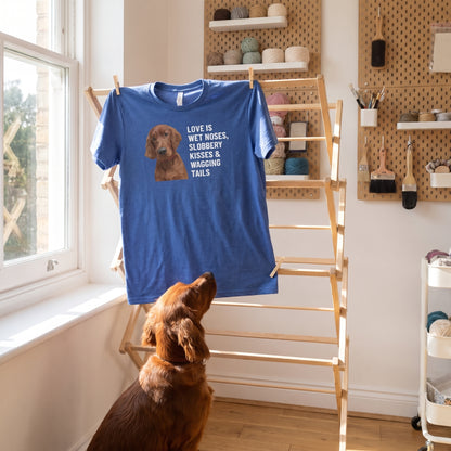 A custom pet t-shirt (Heather True Royal) printed with "Love Is Wet Noses, Slobbery Kisses & Wagging Tails" and an illustration of Finnegan the Irish Setter, in a light-filled nursery-turned-craft room with pegboard shelves, with Finnegan nearby. [ks Colour=Heather True Royal; kind=lifestyle]