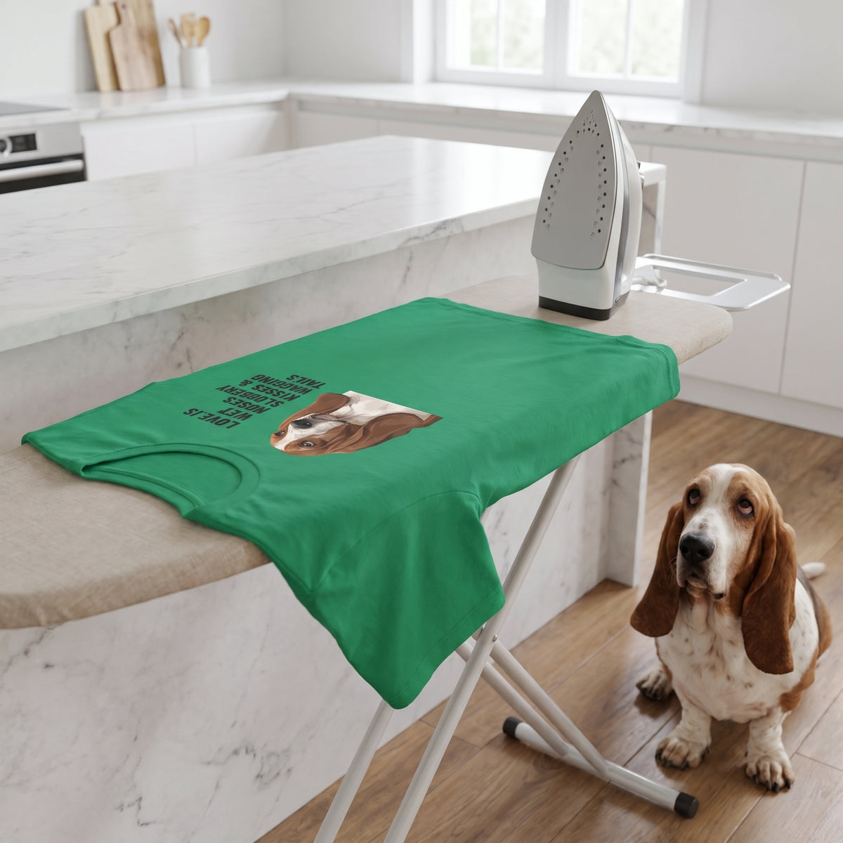 A custom pet t-shirt (Kelly) printed with "Love Is Wet Noses, Slobbery Kisses & Wagging Tails" and an illustration of Bertie the Basset Hound, in a minimalist white kitchen with a marble island, with Bertie nearby. [ks Colour=Kelly; kind=lifestyle]