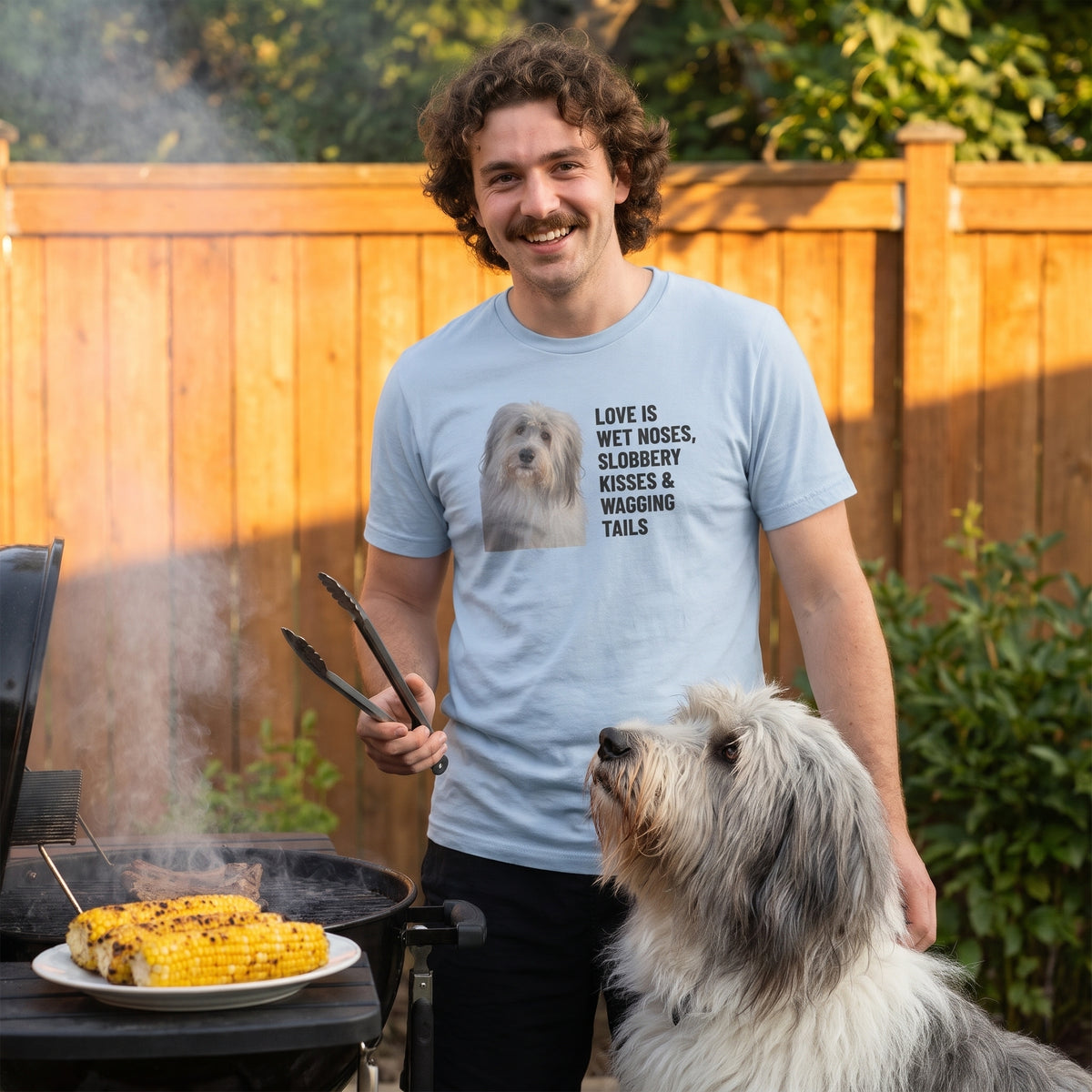 A man in his 20s with curly brown hair and a moustache wearing a custom pet t-shirt (Light Blue) printed with "Love Is Wet Noses, Slobbery Kisses & Wagging Tails" and an illustration of Gandalf the Sheepdog, with Gandalf beside them. [ks Colour=Light Blue; kind=lifestyle]