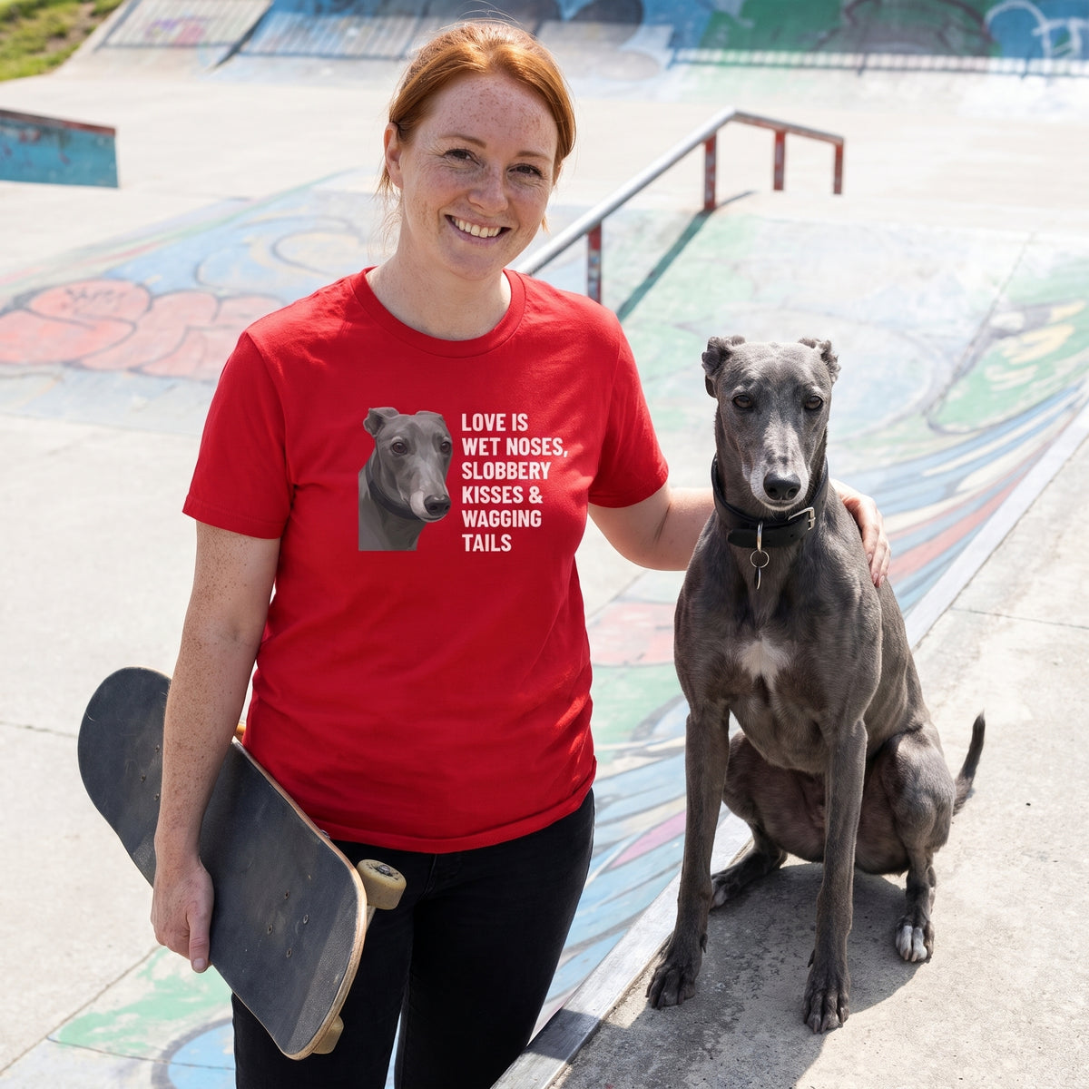 A red-haired freckled woman in her 30s wearing a custom pet t-shirt (Red) printed with "Love Is Wet Noses, Slobbery Kisses & Wagging Tails" and an illustration of Ash the Greyhound, with Ash beside them. [ks Colour=Red; kind=lifestyle]