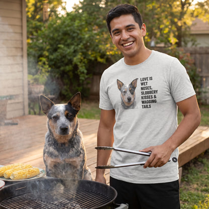 A Latino man in his 20s wearing a custom pet t-shirt (Athletic Heather) printed with "Love Is Wet Noses, Slobbery Kisses & Wagging Tails" and an illustration of Skye the Australian Cattle Dog, with Skye beside them. [ks Colour=Athletic Heather; kind=lifestyle]