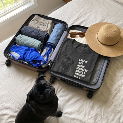 A custom pet t-shirt (Dark Grey Heather) printed with "Love Is Wet Noses, Slobbery Kisses & Wagging Tails" and an illustration of Salem the Pug, at the packed suitcase, with Salem nearby. [ks Colour=Dark Grey Heather; kind=lifestyle]