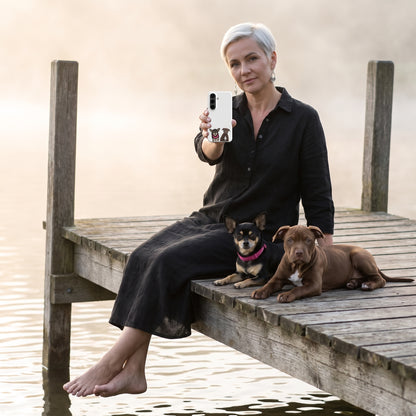 A woman in her 40s with short platinum hair and silver earrings in a black linen dress with a custom pet Galaxy phone case (Snap Matte) featuring an illustration of Taco the Chihuahua & Hershey the Pit Bull, with Taco & Hershey beside them. [ks Style=Snap Matte; kind=lifestyle]